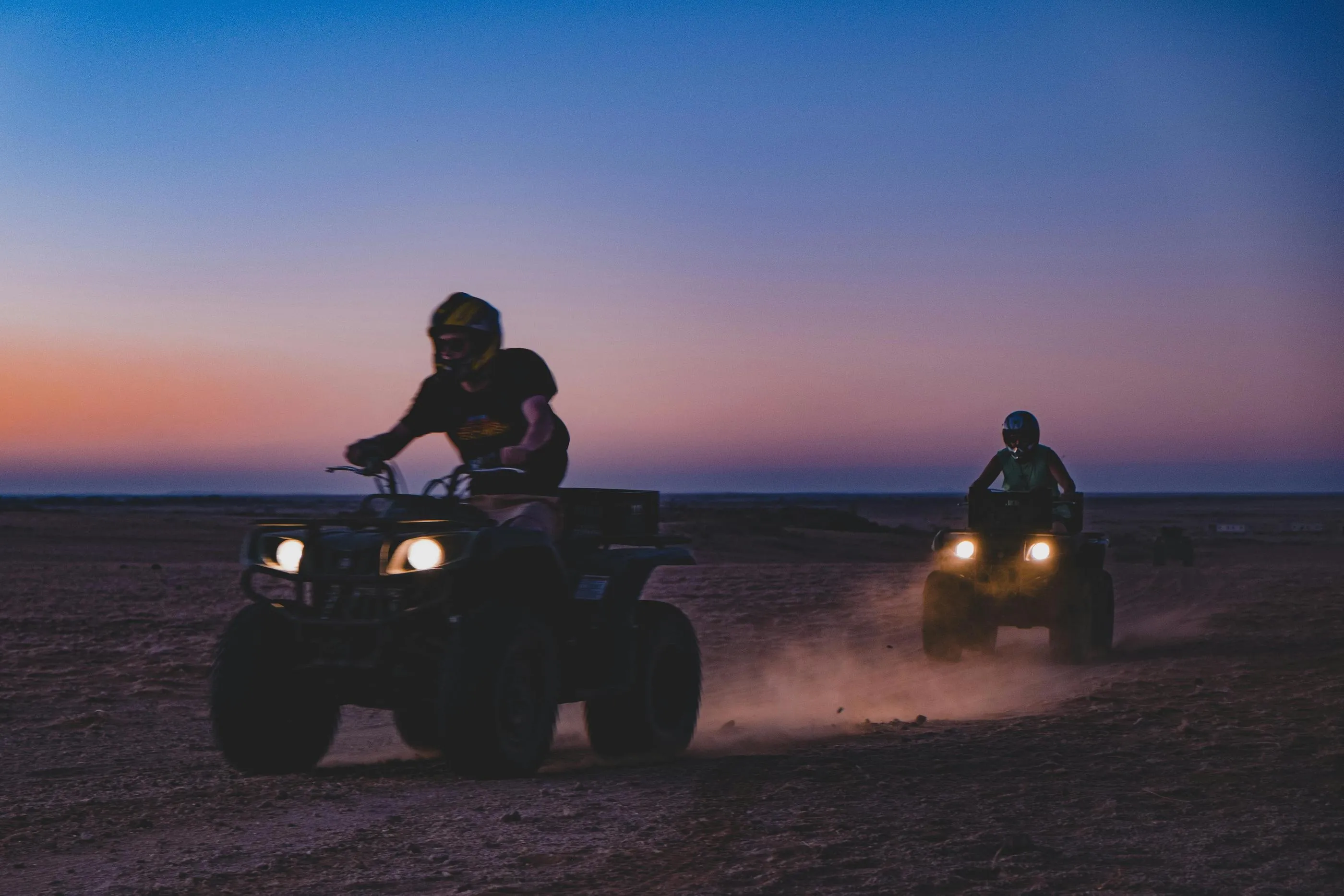 Two people are driving quadricycles in a desert at sunset.