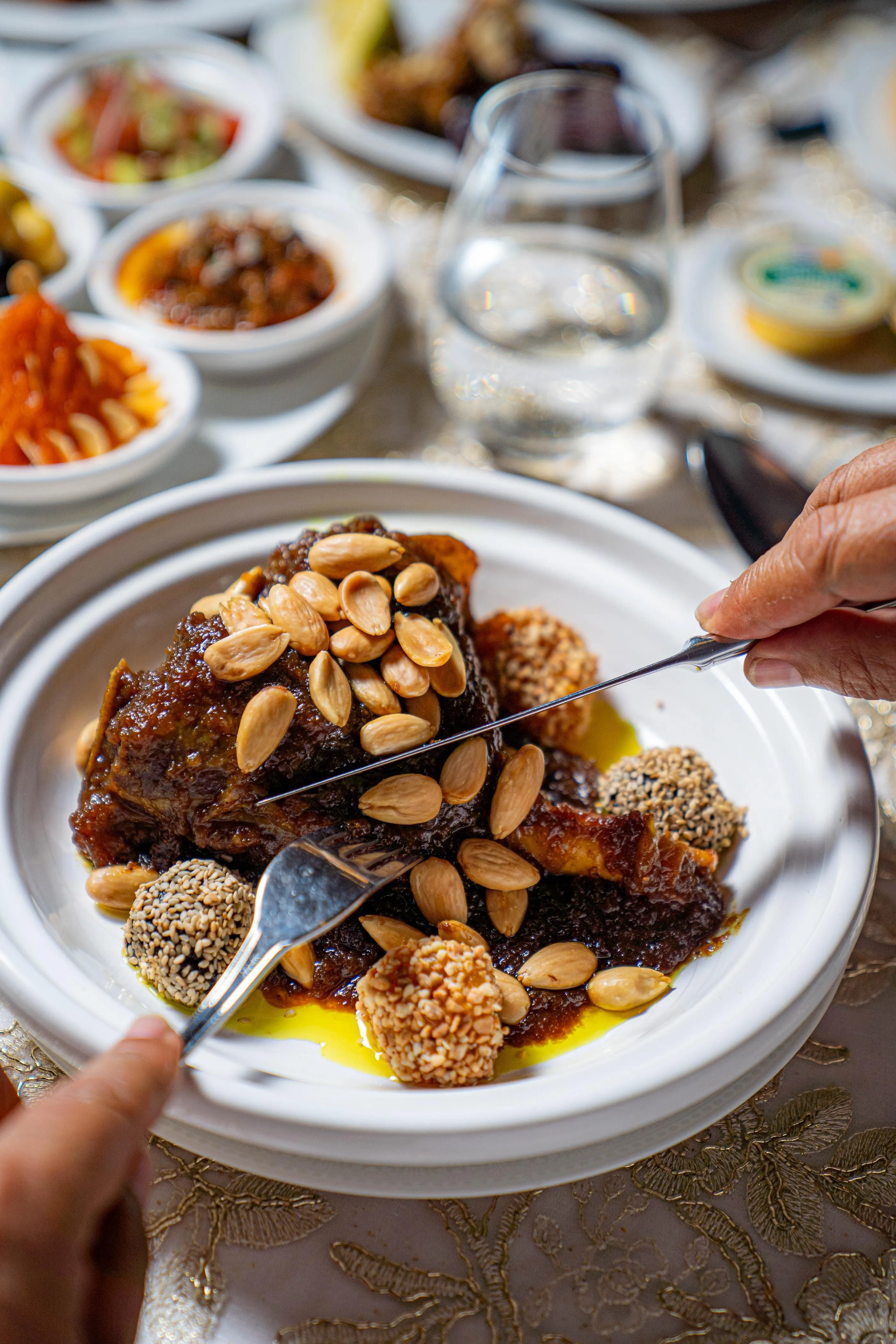Una persona sirve un plato con trozos de carne, almendras y semillas de sésamo sobre una mesa adornada con diversos pequeños platos.