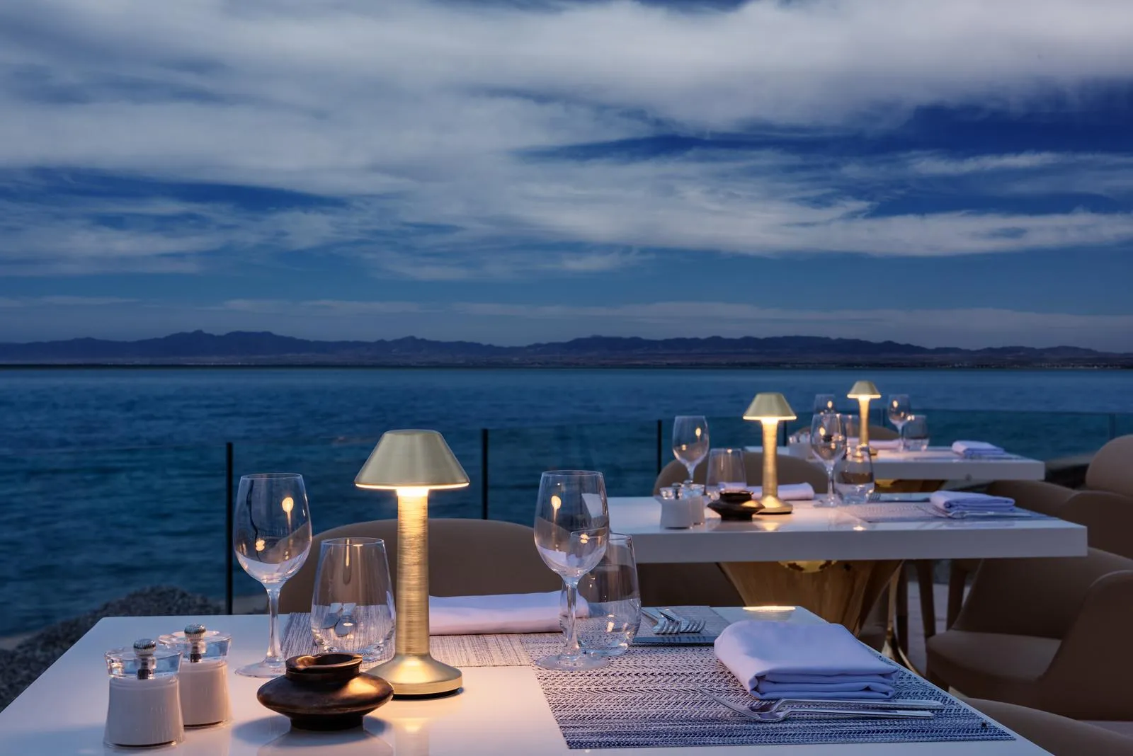 Mesas puestas con vasos y velas en una terraza frente al mar al atardecer