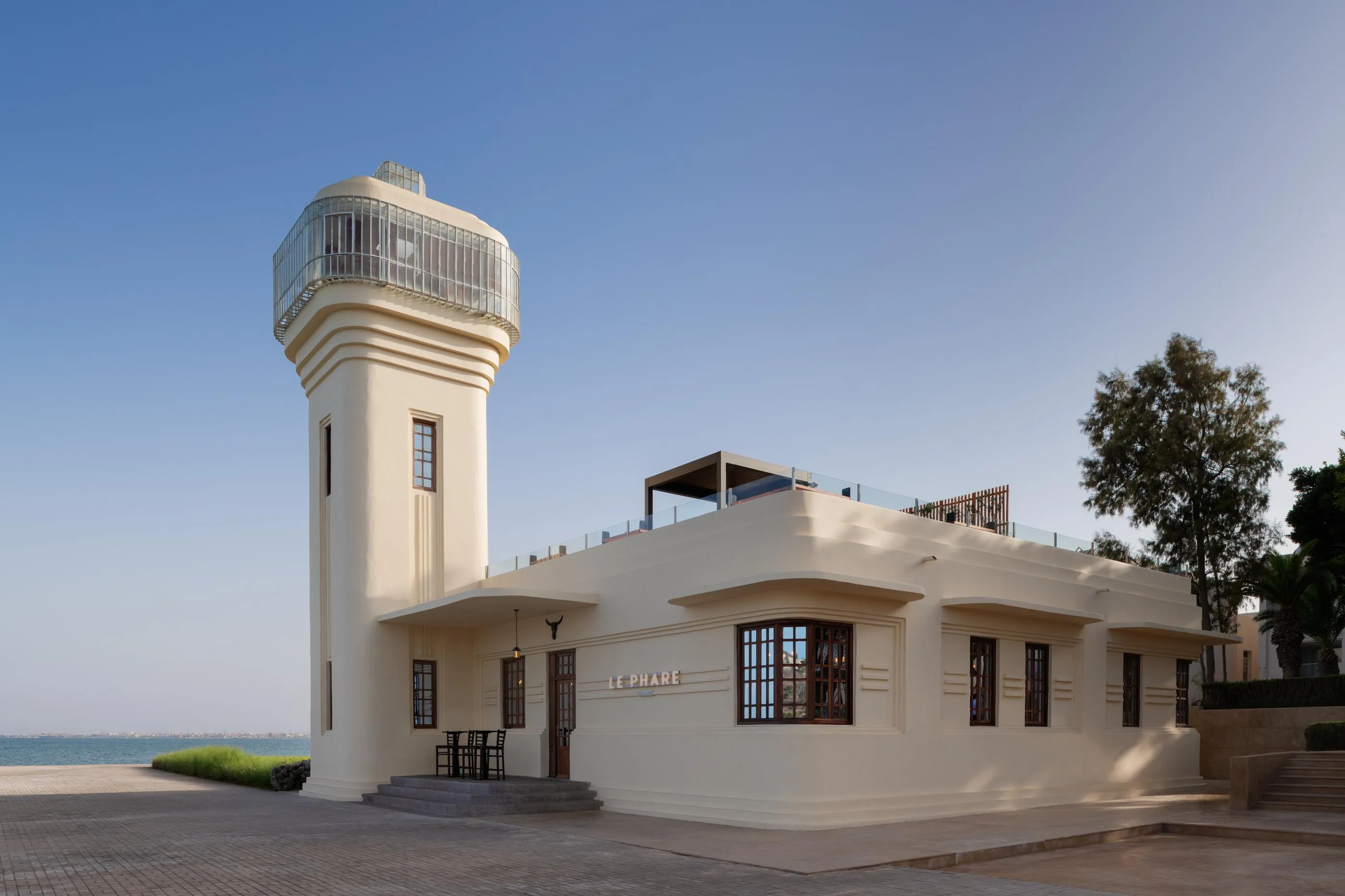 A white lighthouse with a square tower and a glass observation platform, located near the sea, with a low adjacent building bearing the sign "LE PHARE"
