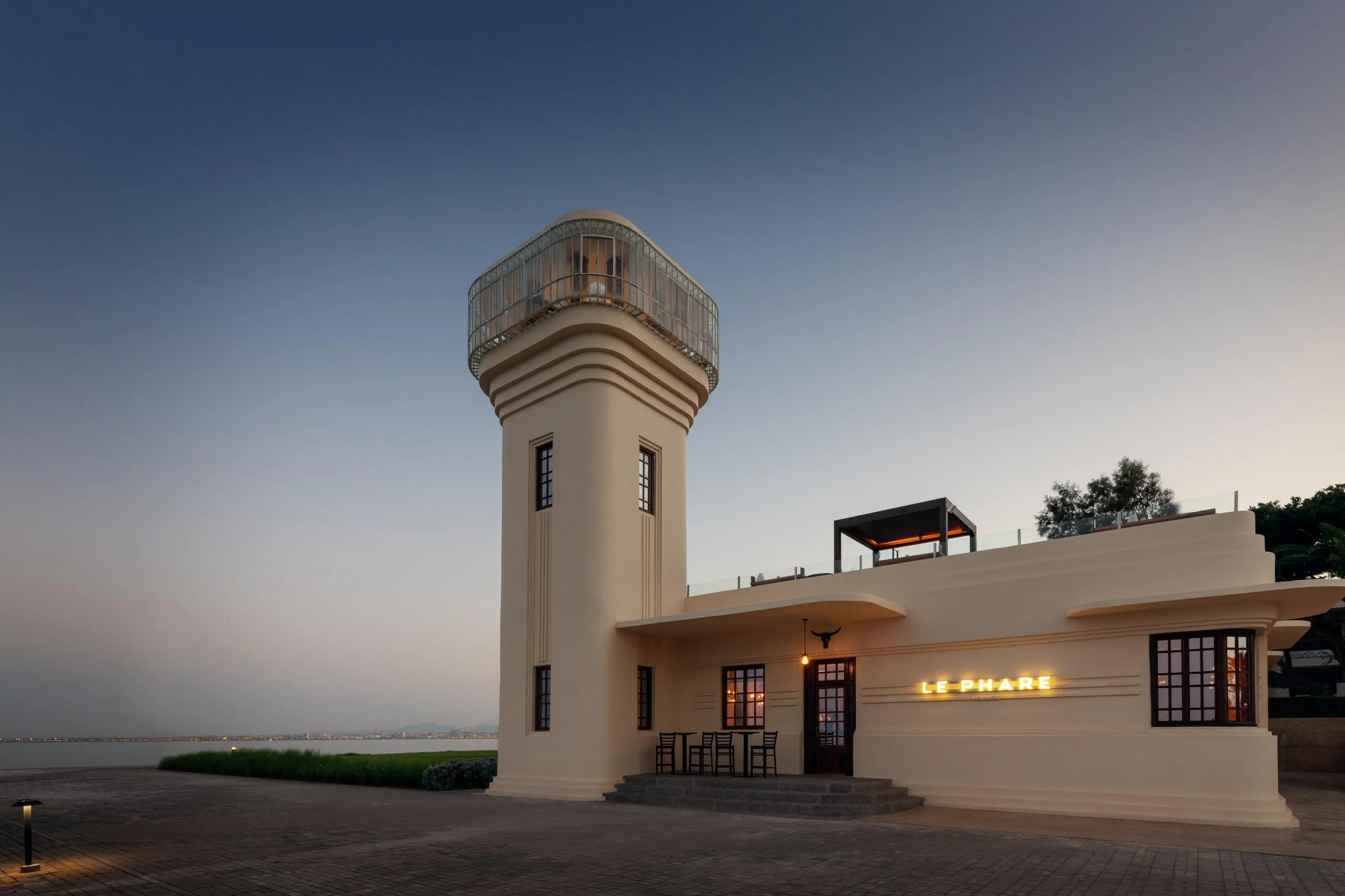 Lighthouse tower with a circular structure and an upper terrace, located near the sea, with an adjacent building bearing the sign "LE PHARE"