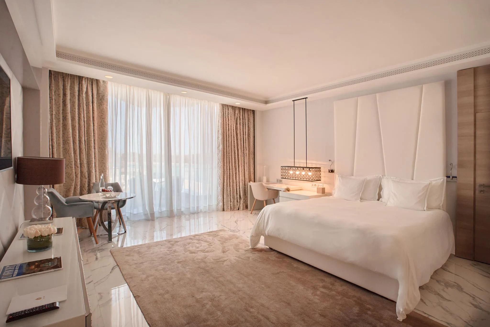 Chambre d'hôtel avec un grand lit blanc, une table de chevet avec une lampe, un coin salon près d'une grande fenêtre aux rideaux ouverts