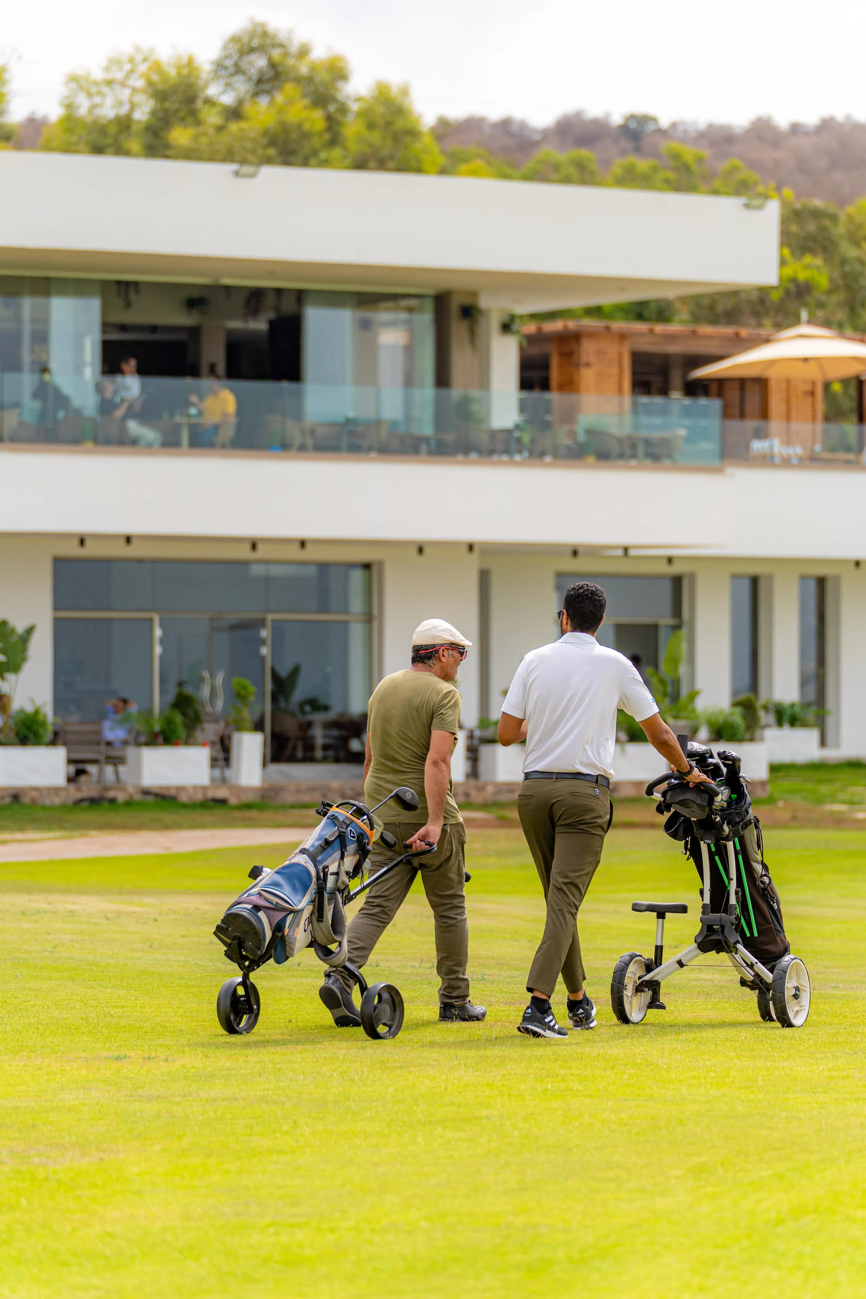 Dos hombres caminan por un campo de golf con bolsas de palos, frente a un edificio moderno con grandes ventanales.