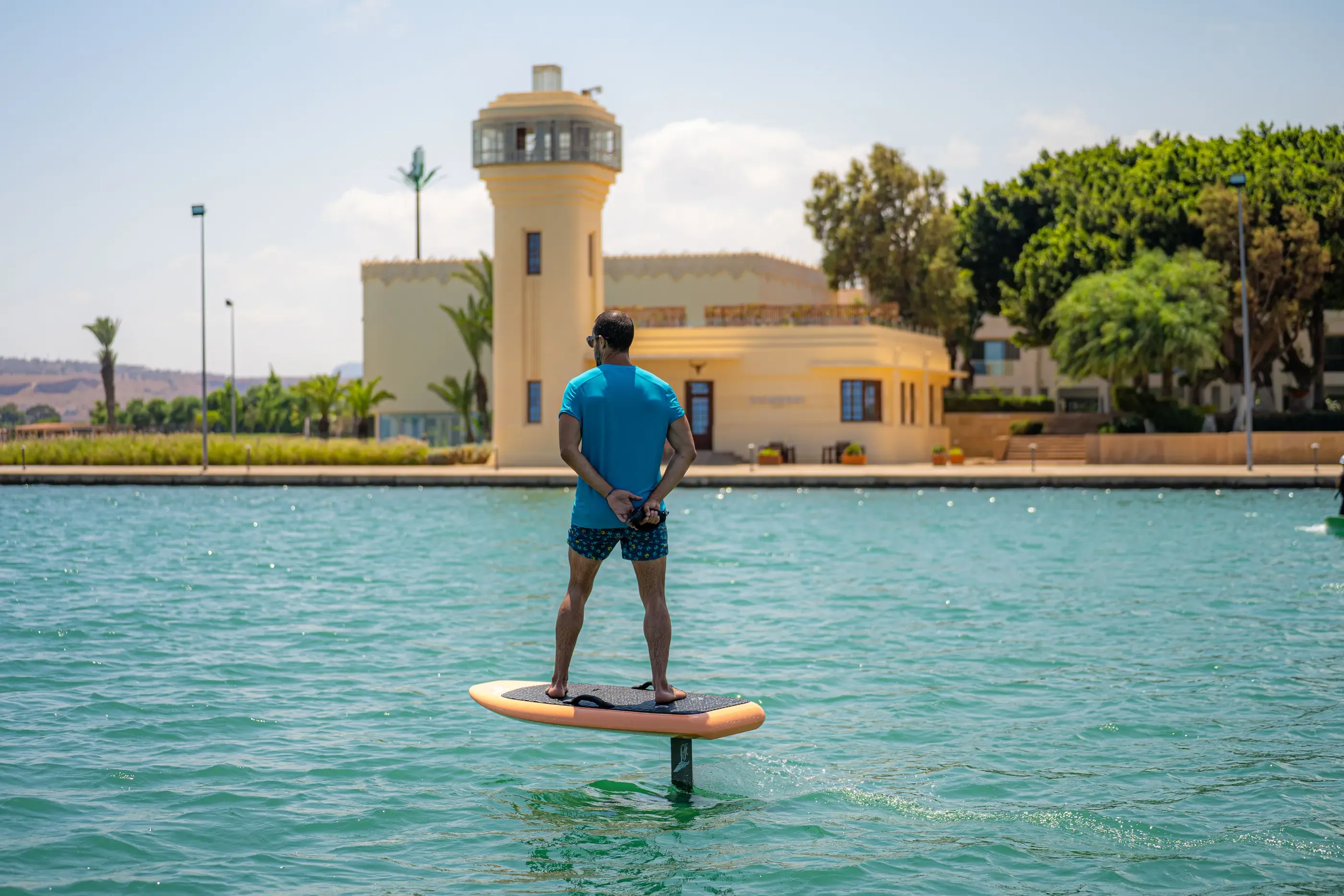 A man standing on a paddleboard in a body of water, with a yellow building in the background.