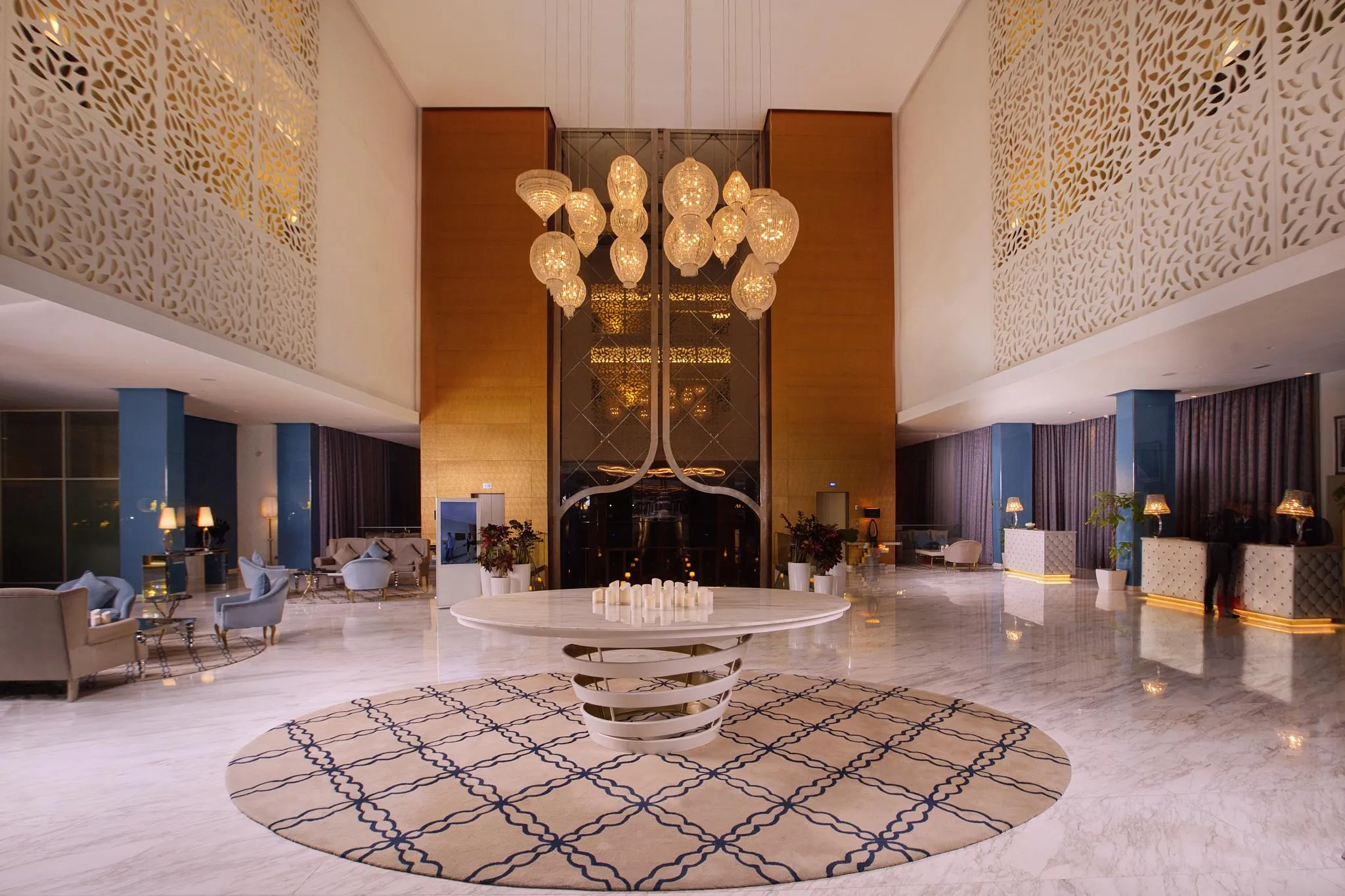 Luxurious hotel lobby with a large chandelier, sofas, coffee tables, and walls decorated with geometric patterns.