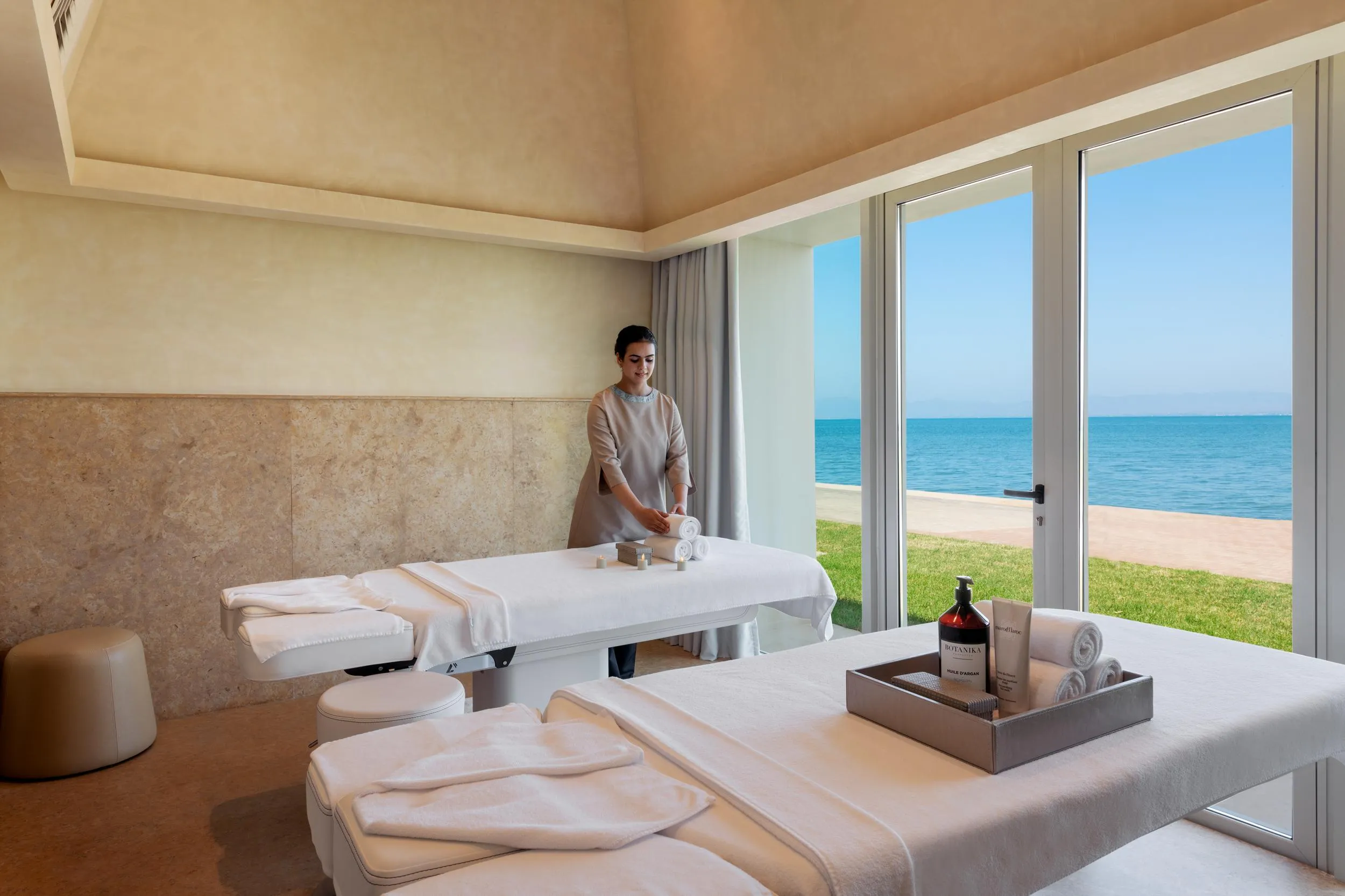 Una persona con ropa de trabajo prepara una mesa de masaje en una sala con vista al mar.