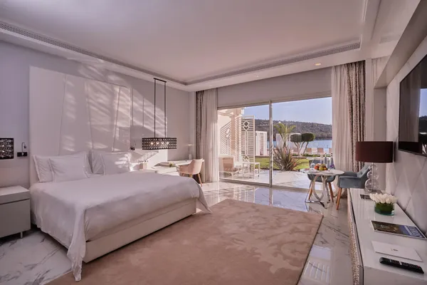 Habitación de hotel moderna con una cama grande, una mesa y sillas cerca de una puerta de cristal que da a una terraza con vistas al agua