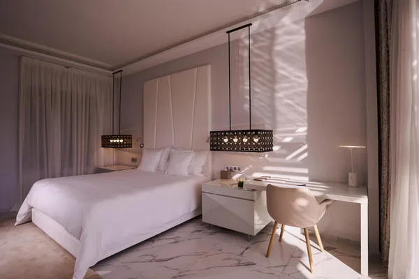 Chambre avec un lit double blanc, une table de chevet et une chaise près d'une fenêtre aux rideaux translucides