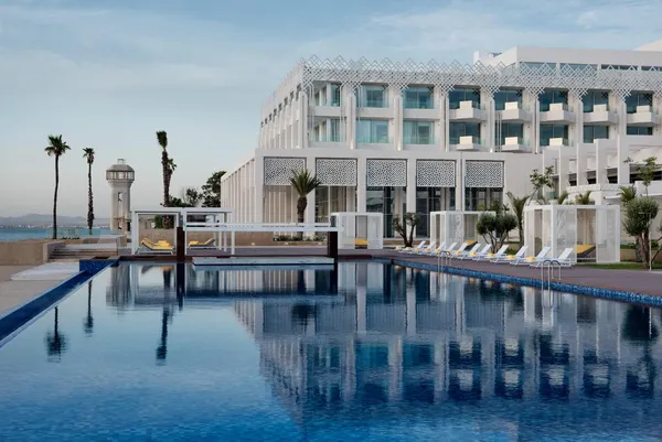 Piscina de borde infinito frente a un hotel moderno con tumbonas y palmeras, vista al mar de fondo