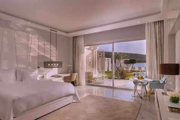Habitación lujosa con cama blanca, escritorio y vistas a una terraza y un paisaje exterior desde grandes ventanales.