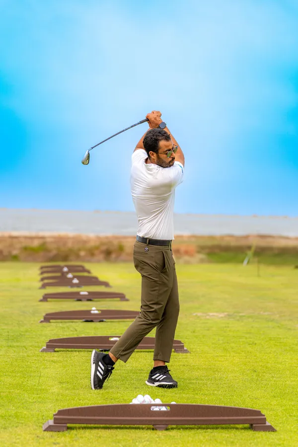 Un hombre con polo blanco y pantalón verde golpea pelotas de golf en un campo de práctica al aire libre.