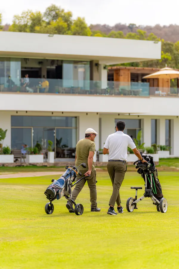 Dos hombres caminan por un campo de golf con bolsas de palos, frente a un edificio moderno con grandes ventanales.