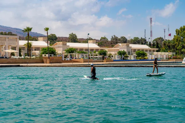 Dos personas hacen paddle en una extensión de agua turquesa frente a un complejo de edificios blancos y palmeras.