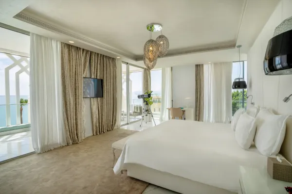 Chambre moderne avec un grand lit blanc, des fenêtres donnant sur la mer, des rideaux, un téléviseur mural et un éclairage suspendu