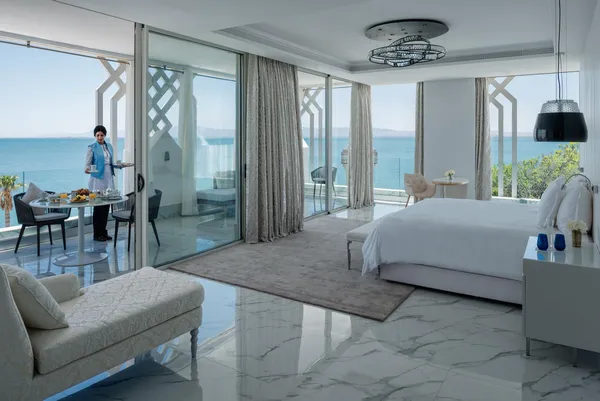 Luxurious hotel room with sea view, a bed, a sofa, a dining table and a person serving a tray