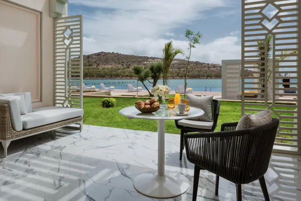 Terraza con mesa, sillas y sofá que da a un césped y un lago con tumbonas y montañas de fondo